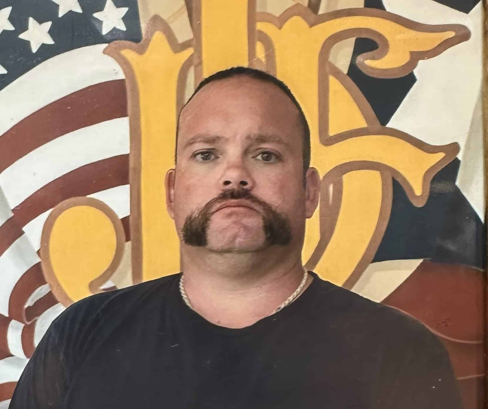 A man with a mustache in front of a mural of the American flag and the number 16.