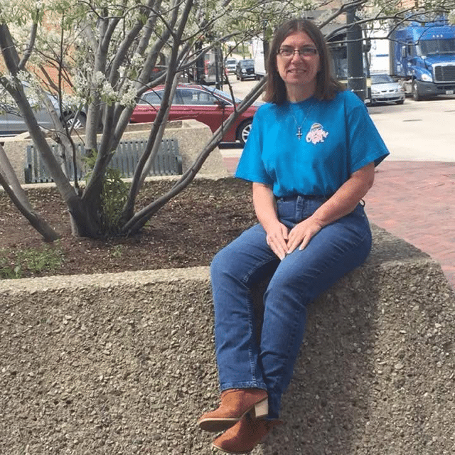 A woman with glasses wearing a blue t-shirt and jeans sits on a concrete ledge next to a flowering tree.